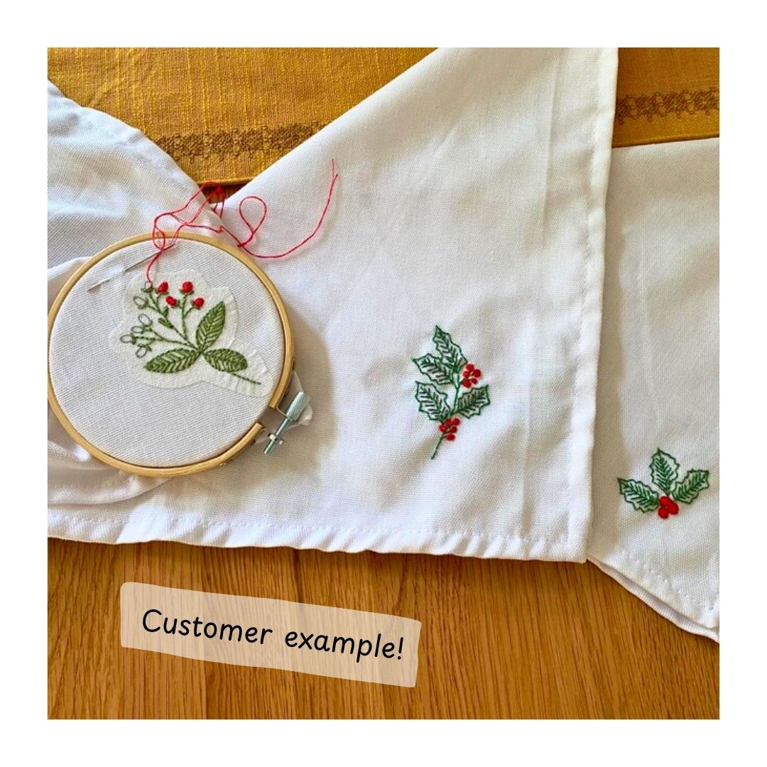 Embroidery hoop with festive design on white napkins with embroidered patterns on a wooden surface.