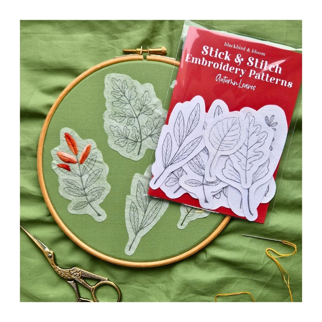Embroidery hoop with leaf patterns, scissors, and 'Stick & Stitch' embroidery pattern book on a green fabric background.