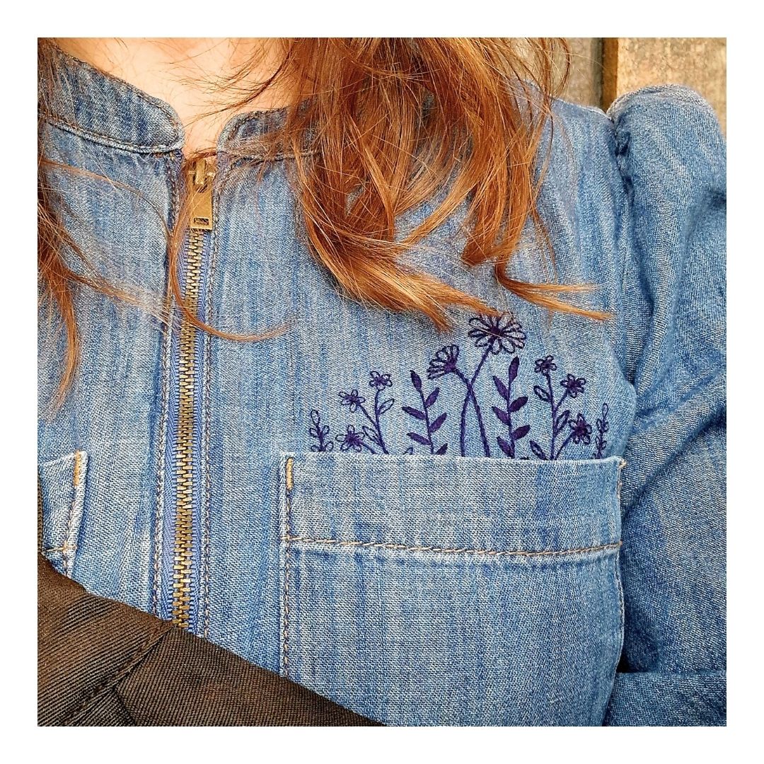 A close-up of a person wearing a light blue denim zip-up dress with dark purple floral embroidery on the chest pocket and a gold zipper.