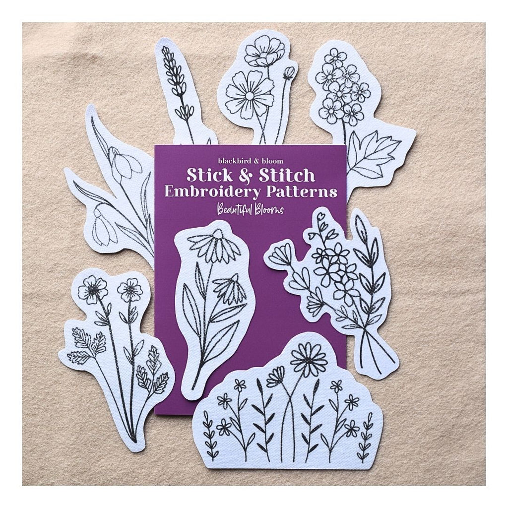 A set of floral embroidery patterns arranged around a purple booklet on a beige textured background. The booklet reads 'blackbird & bloom' Stick & Stitch Embroidery Patterns Beautiful Blooms'.