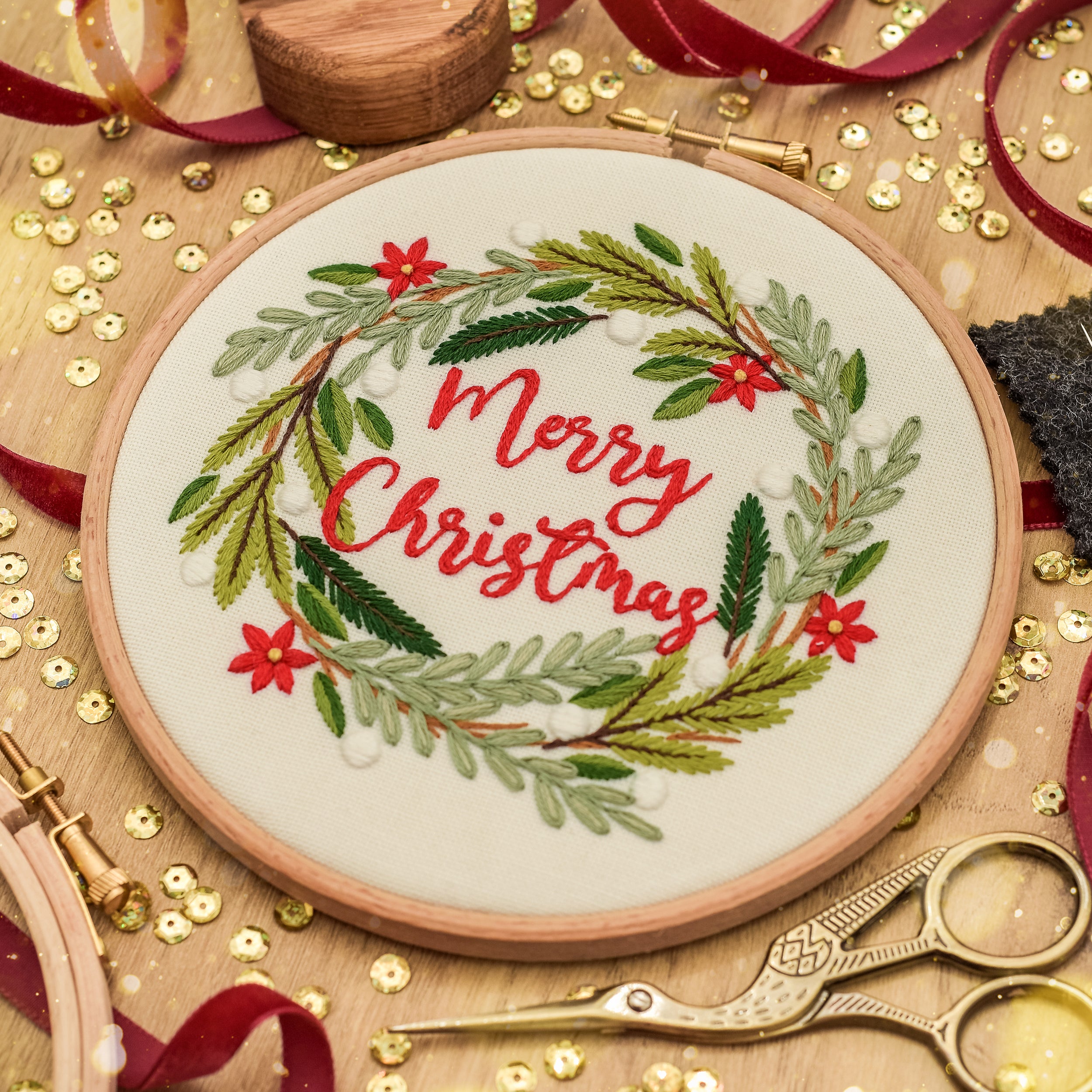 A round wooden embroidery hoop containing a festive 'Merry Christmas' stitched design with green pine branches, red flowers, and red lettering, displayed on a wooden surface with gold sequins, ribbon, scissors, and other craft tools.