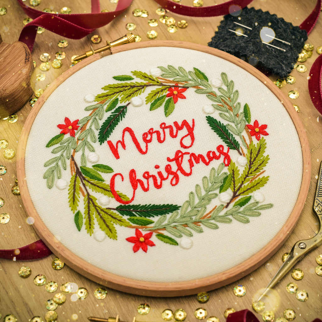 A round wooden embroidery hoop holding a white fabric with a festive Christmas wreath design. The wreath is made of green and light green embroidered leaves, accented with small red flowers and white pearl-like beads, with 'Merry Christmas' stitched in red cursive script in the center.