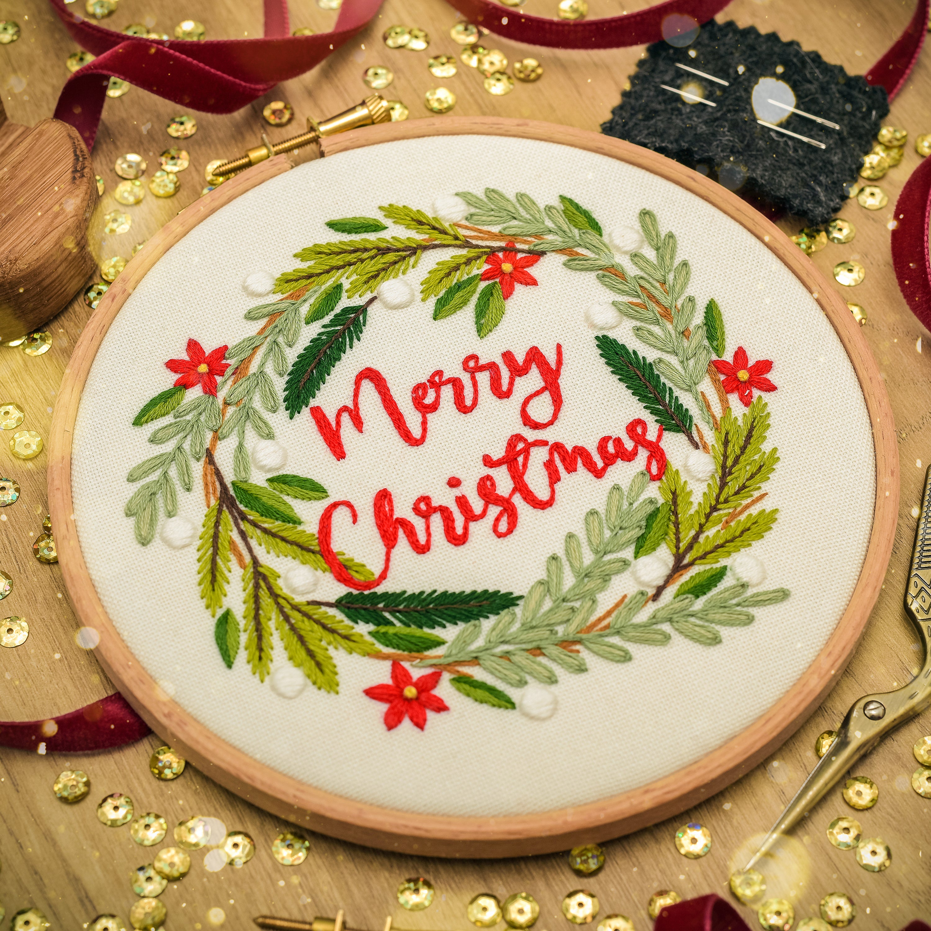 A round wooden embroidery hoop holding a white fabric with a festive Christmas wreath design. The wreath is made of green and light green embroidered leaves, accented with small red flowers and white pearl-like beads, with 'Merry Christmas' stitched in red cursive script in the center.