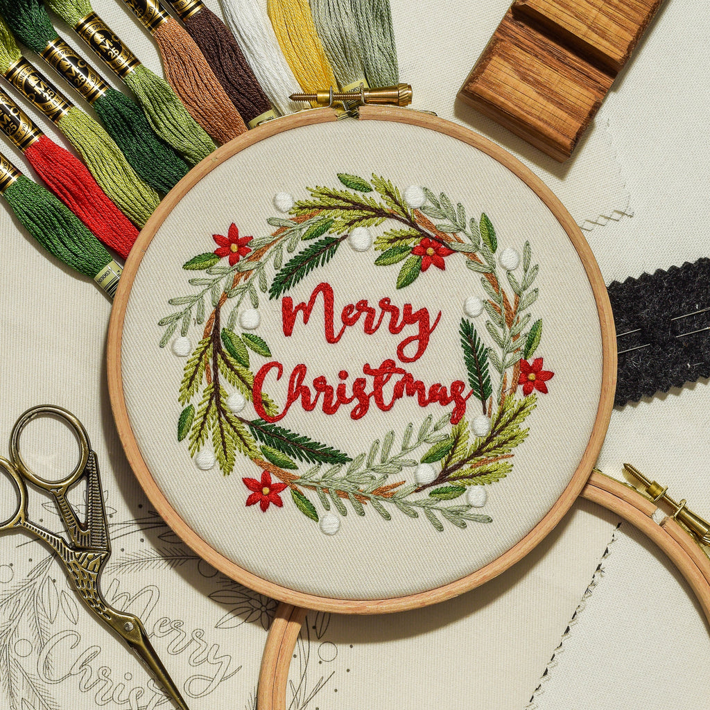 An embroidery hoop displays a completed 'Merry Christmas' wreath design in red and green thread, surrounded by embroidery floss, scissors, wooden blocks, and pattern paper on a light background.