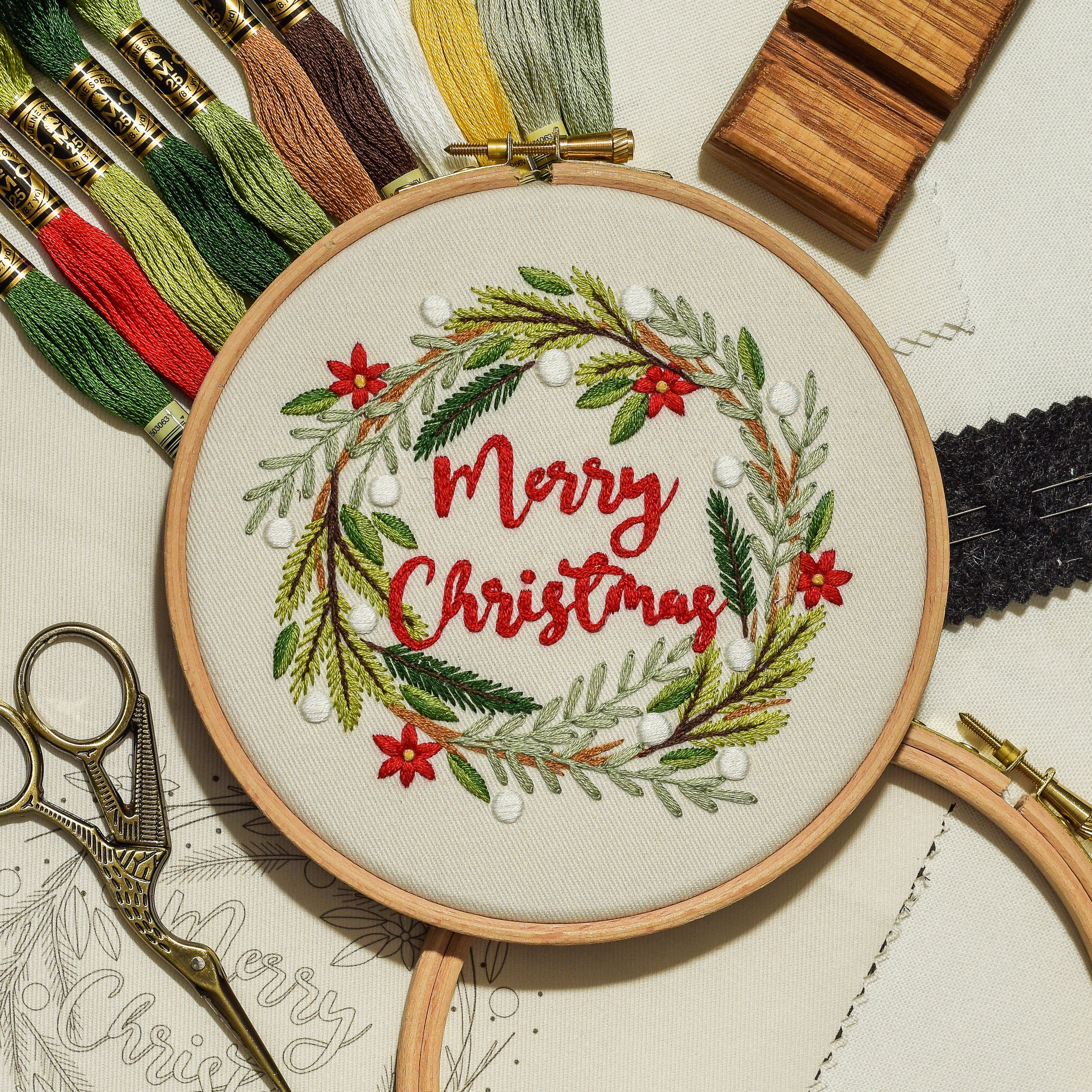 An embroidery hoop displays a completed 'Merry Christmas' wreath design in red and green thread, surrounded by embroidery floss, scissors, wooden blocks, and pattern paper on a light background.