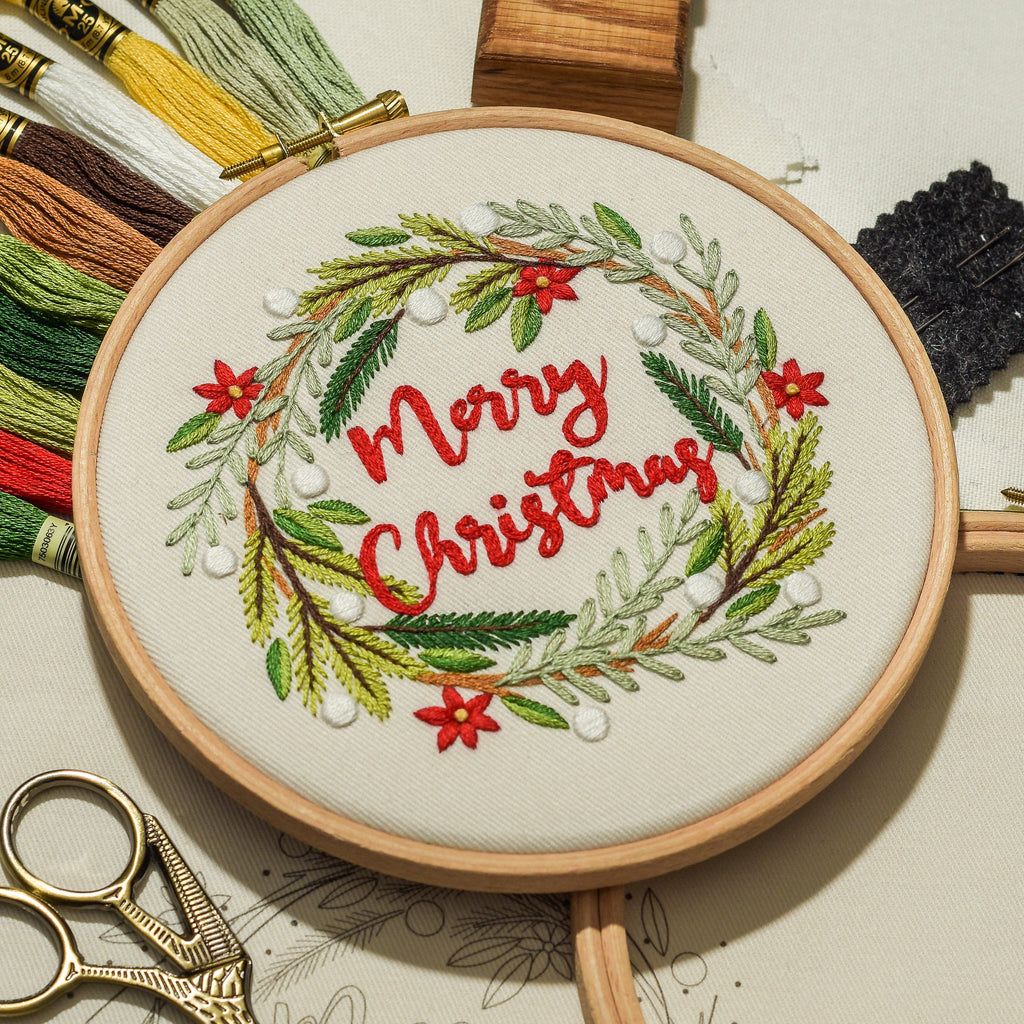 An embroidery hoop displays a festive Christmas wreath design with red 'Merry Christmas' lettering, surrounded by green leaves and red flowers. The hoop is styled with embroidery floss, gold scissors, and wooden blocks on a light beige background.