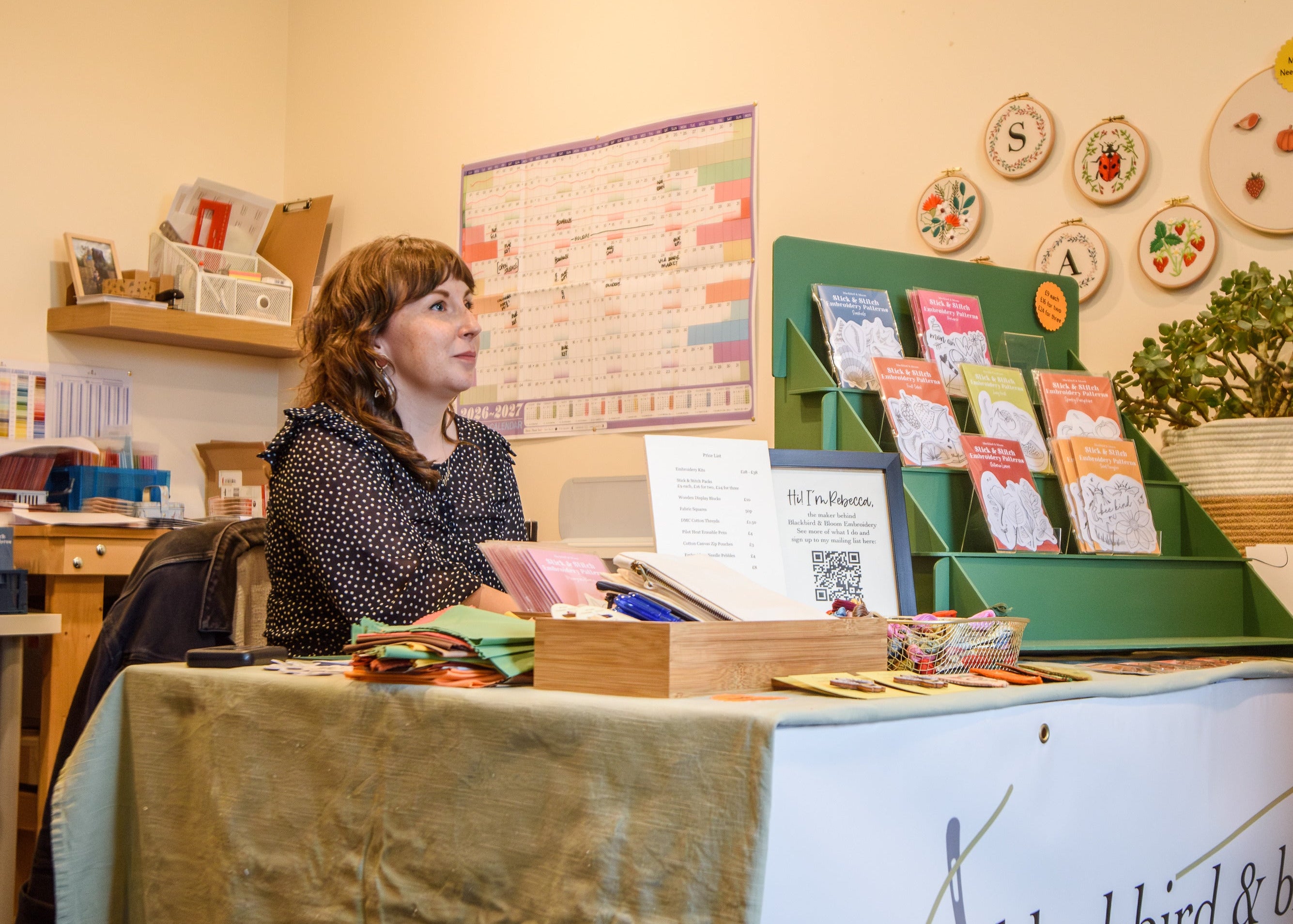 A woman sits behind a display table at an open studios event. The table has a white front banner reading 'blackbird & bloom embroidery' and displays books, cards, and small fabric and craft items.