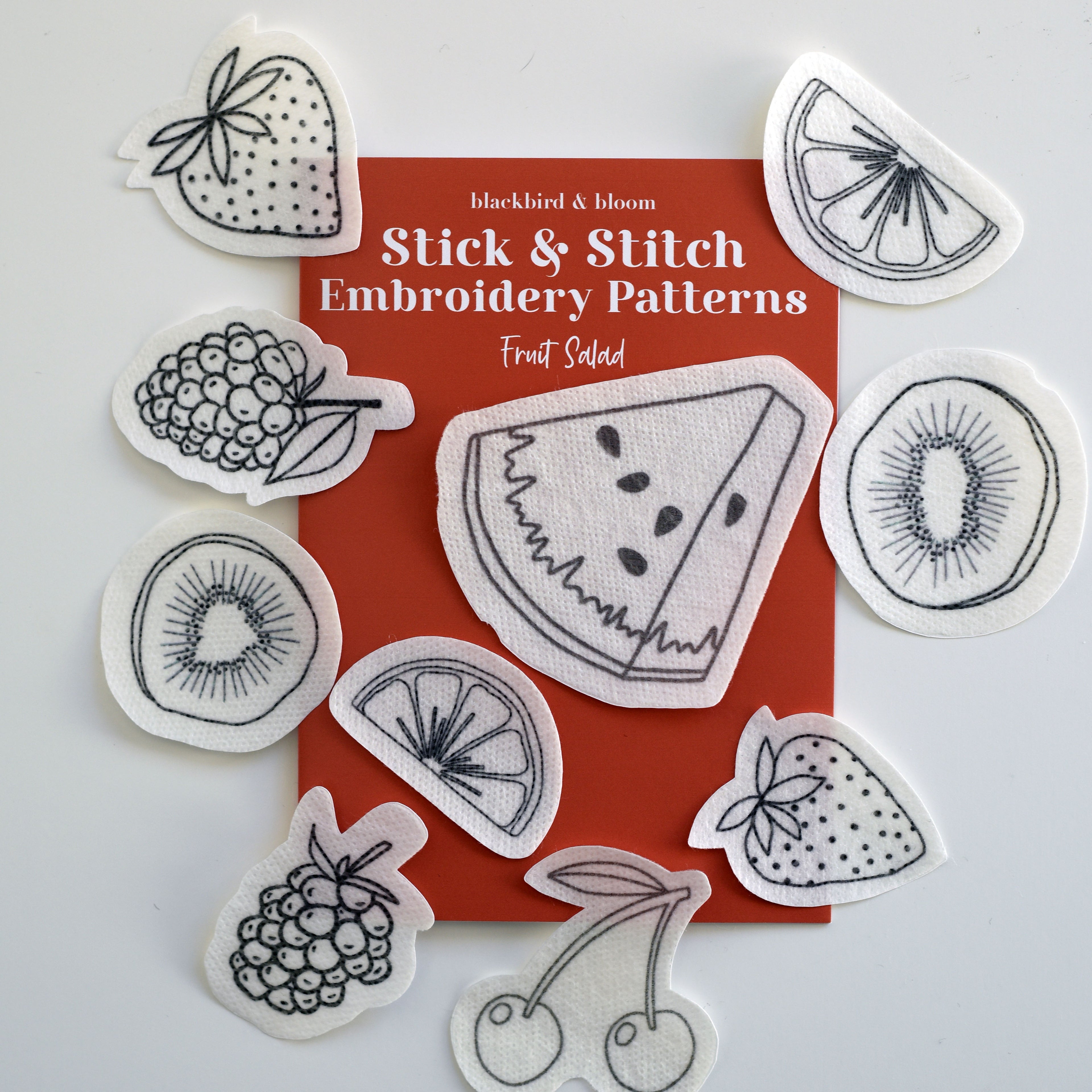 Various embroidery patterns of fruit salad designs scattered around an orange guide card with the title 'Stick & Stitch Embroidery Patterns' by Blackbird & Bloom.