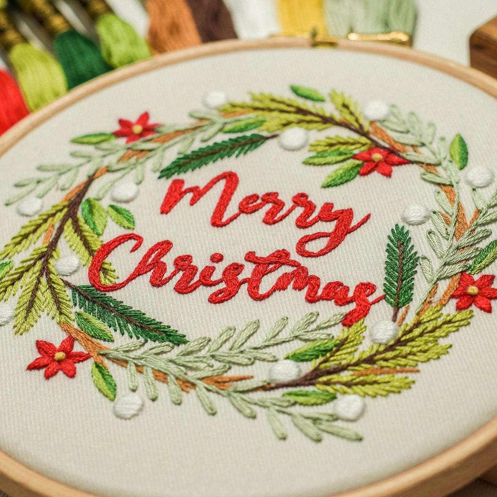 A close-up of a wooden embroidery hoop holding a festive Christmas wreath design with red embroidered 'Merry Christmas' text, green pine branches, white berries, and red poinsettias on cream fabric. Colorful embroidery floss skeins are blurred in the background.